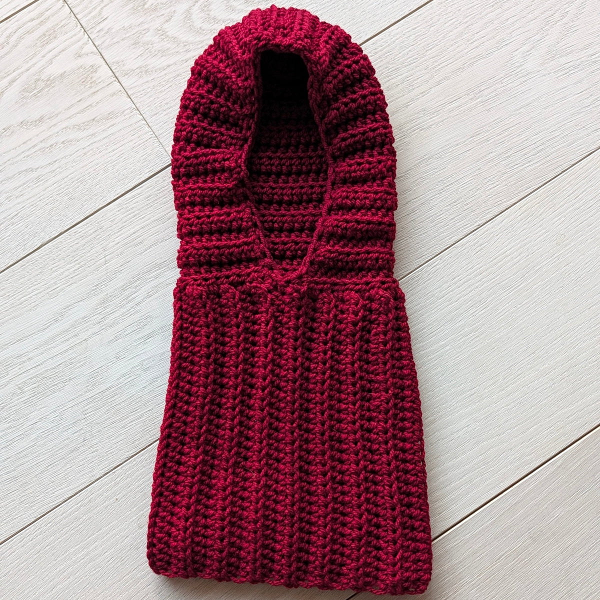 Burgundy chunky crochet balaclava laid flat on a light wooden floor, showing the ribbed turtleneck, textured hood and oval face opening.