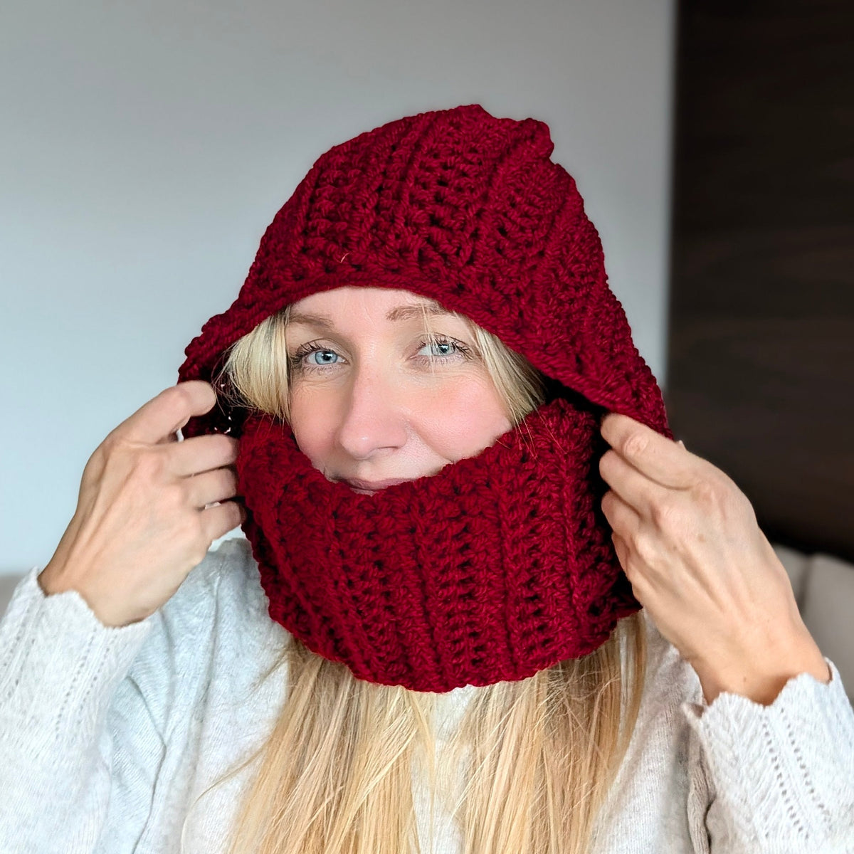 Woman adjusting a burgundy chunky crochet balaclava hood with both hands, showing the textured hood and ribbed turtleneck pulled up over her mouth.