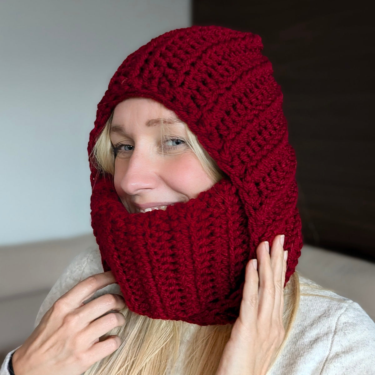 Smiling woman wearing a burgundy chunky crochet balaclava, holding the sides of the raised hood and face-covering ribbed turtleneck.