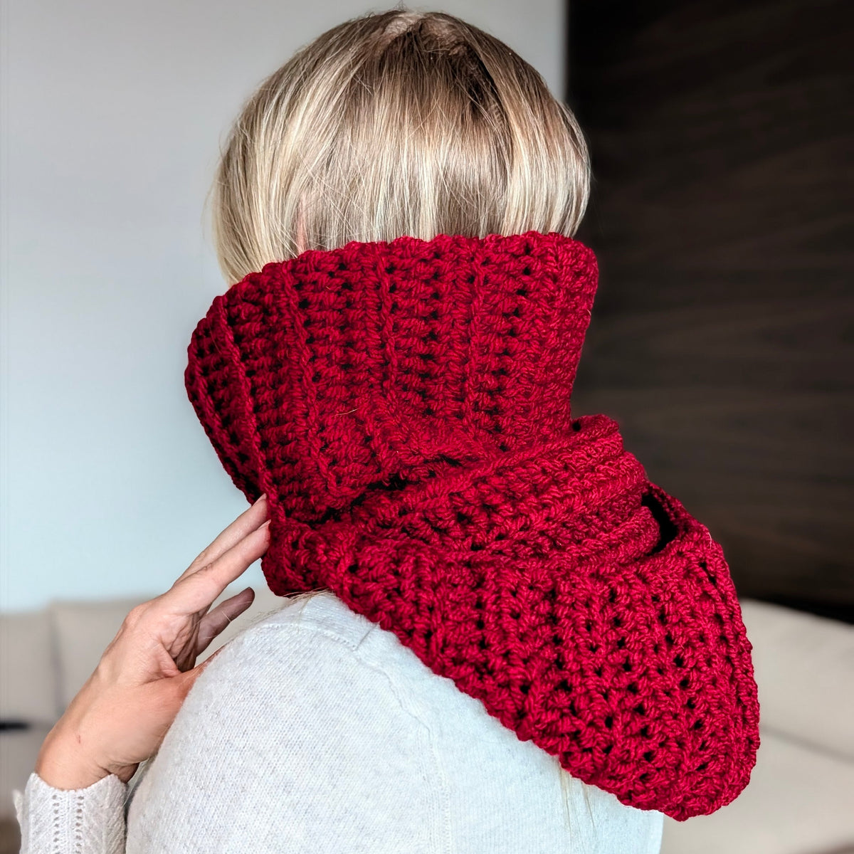 Back view of a woman wearing a burgundy chunky crochet balaclava with the hood folded down, showing the ribbed turtleneck and textured hood resting on her shoulders.