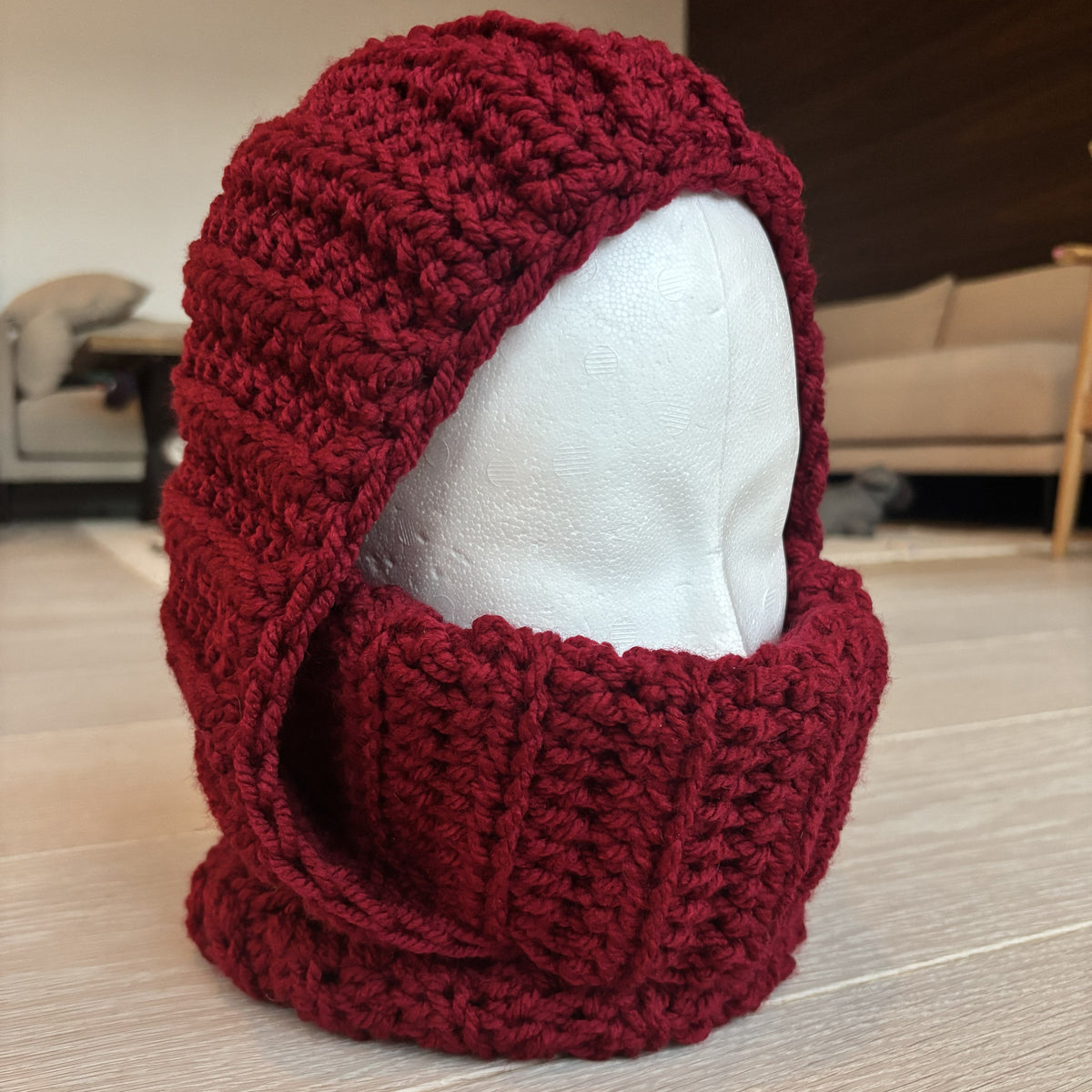 Front three-quarter view of a burgundy crocheted balaclava displayed on a white mannequin head, showing the hood opening, raised ribbed neck and thick stitches.