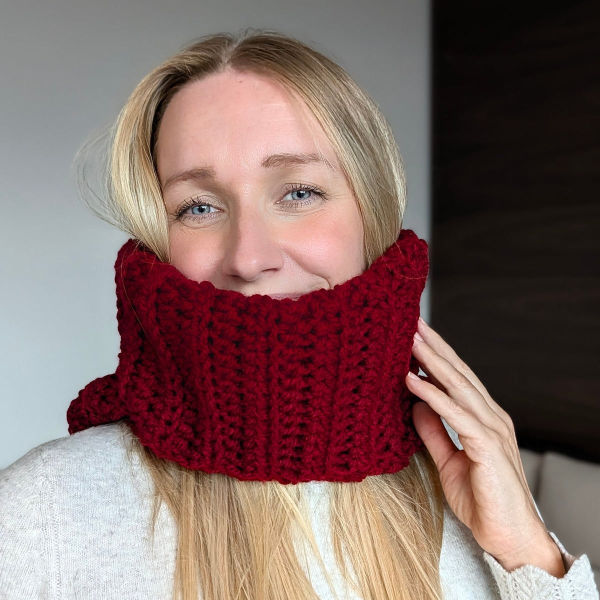 Woman smiling with the burgundy ribbed neck section of a crochet balaclava pulled over her mouth, with the hood resting behind her shoulders.