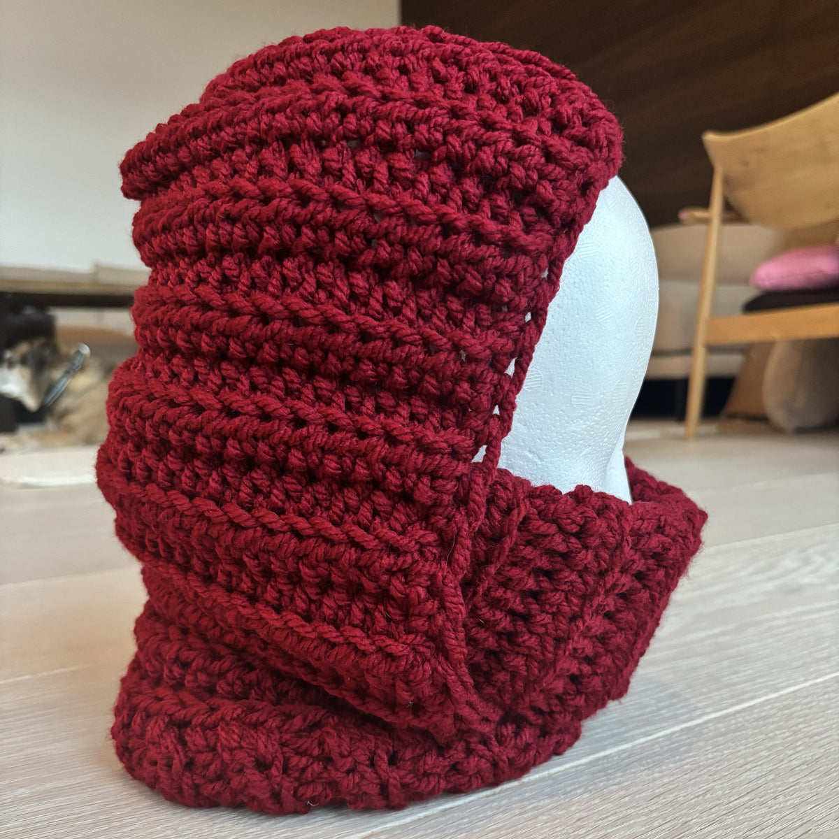 Side view of a burgundy chunky crochet balaclava on a white mannequin head, showing the horizontal hood rows, ribbed turtleneck and layered construction.