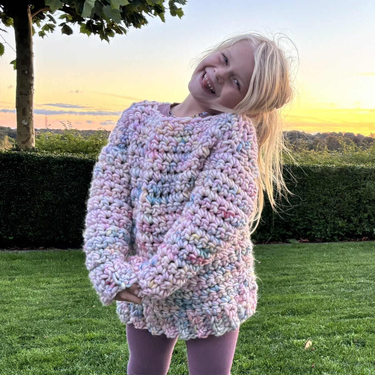 Back view of a handmade chunky crochet kids sweater in pastel pink, blue, lilac and mint, with textured stitches and a ribbed lower edge.