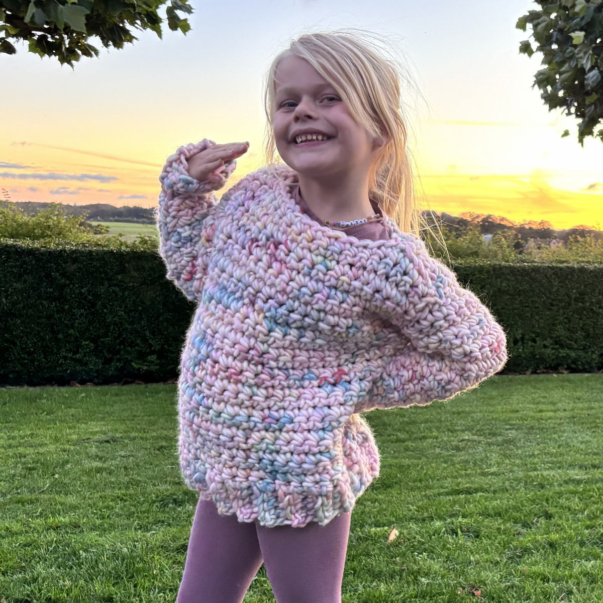 Young girl modeling a chunky pastel crochet sweater with ribbed hem and long sleeves, shown in two playful front poses outdoors.