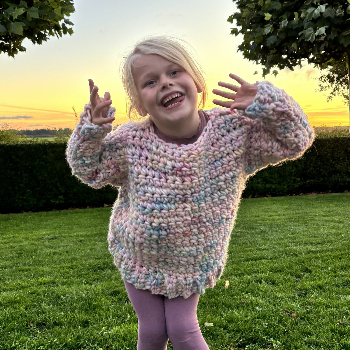 Back view of a handmade chunky crochet kids sweater in pastel pink, blue, lilac and mint, with textured stitches and a ribbed lower edge.