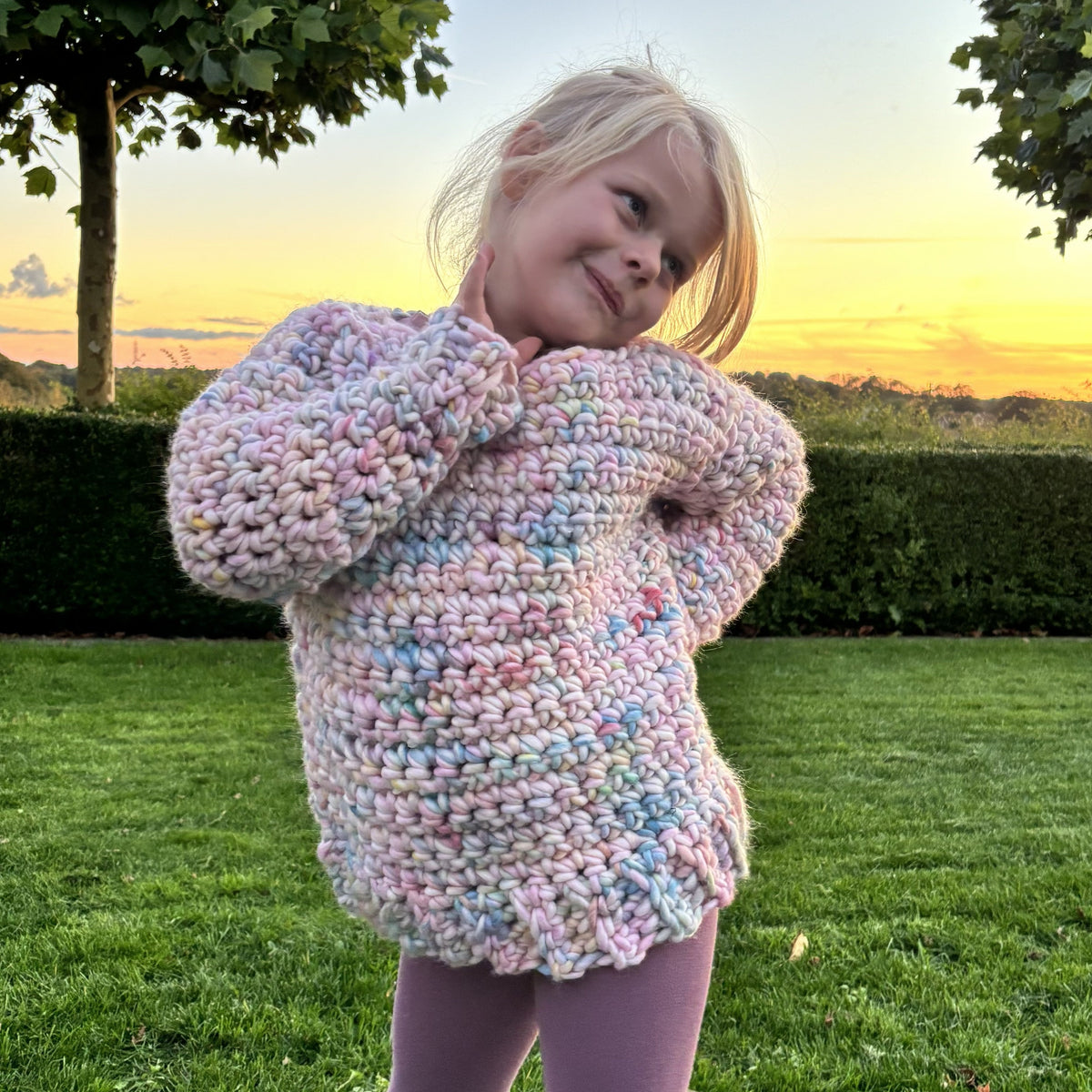 Young girl modeling a chunky pastel crochet sweater with ribbed hem and long sleeves, shown in two playful front poses outdoors.