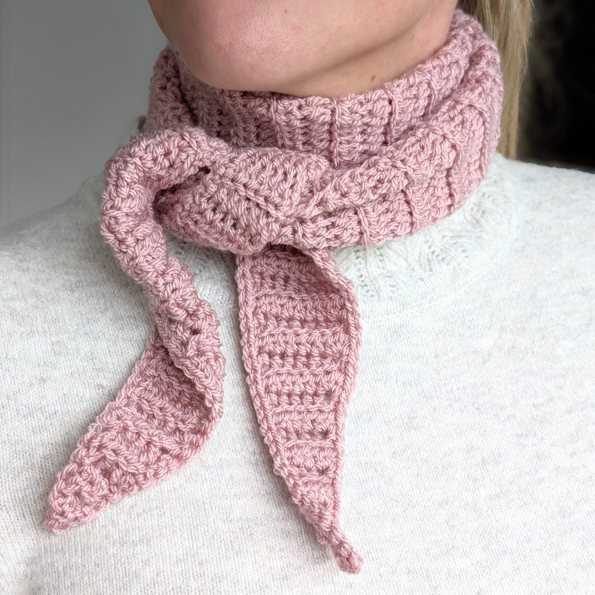 Close-up of a dusty pink crochet Sophie-style neck scarf showing raised ribbed stitches, a wrapped neck section and two pointed ends.