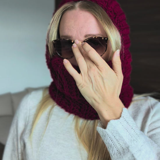 A model showcases the finished burgundy crochet balaclava while wearing the hood, highlighting the chunky stitch texture, face opening and ribbed neck covering.