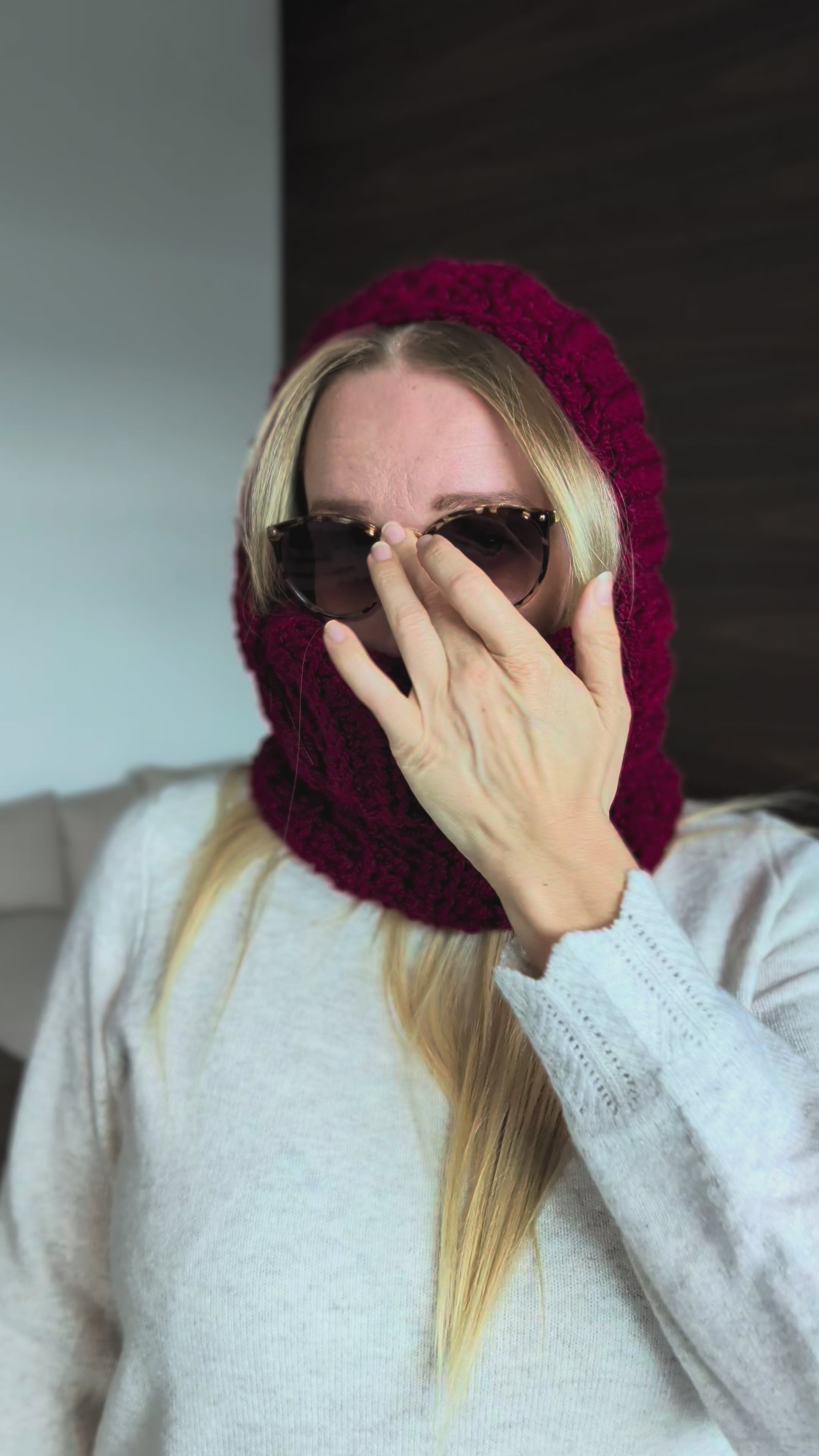 A model showcases the finished burgundy crochet balaclava while wearing the hood, highlighting the chunky stitch texture, face opening and ribbed neck covering.