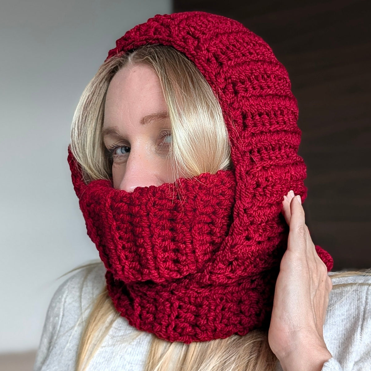 Woman wearing a dark red chunky crochet balaclava with a ribbed turtleneck pulled up over her mouth and a textured hood framing her face.