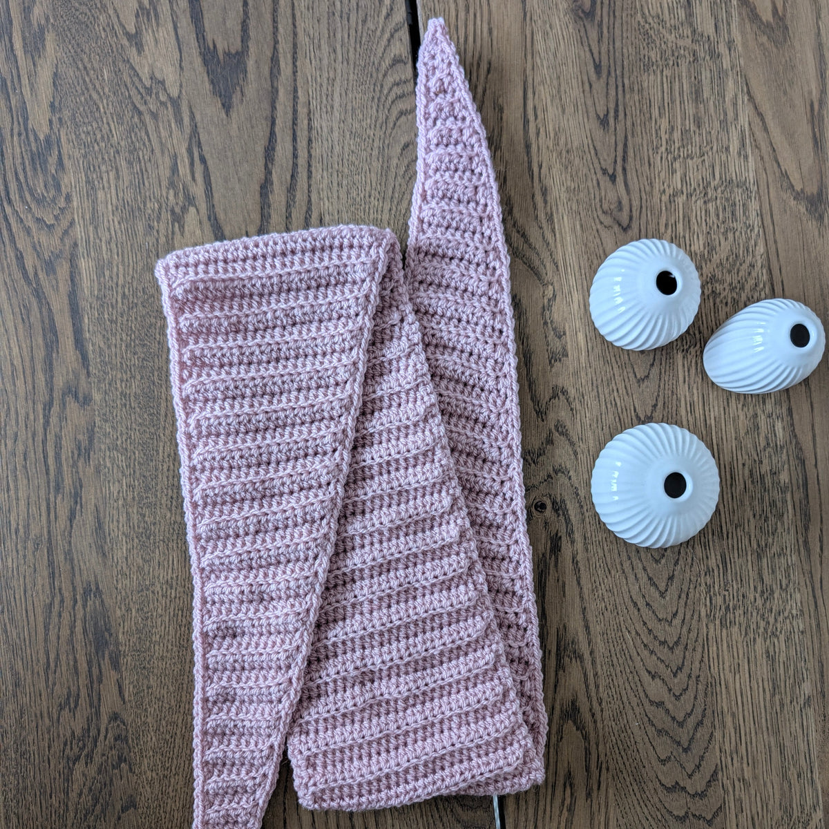 Dusty pink ribbed crochet Sophie Scarf arranged on a wooden tabletop with both pointed ends visible beside three small white vases.