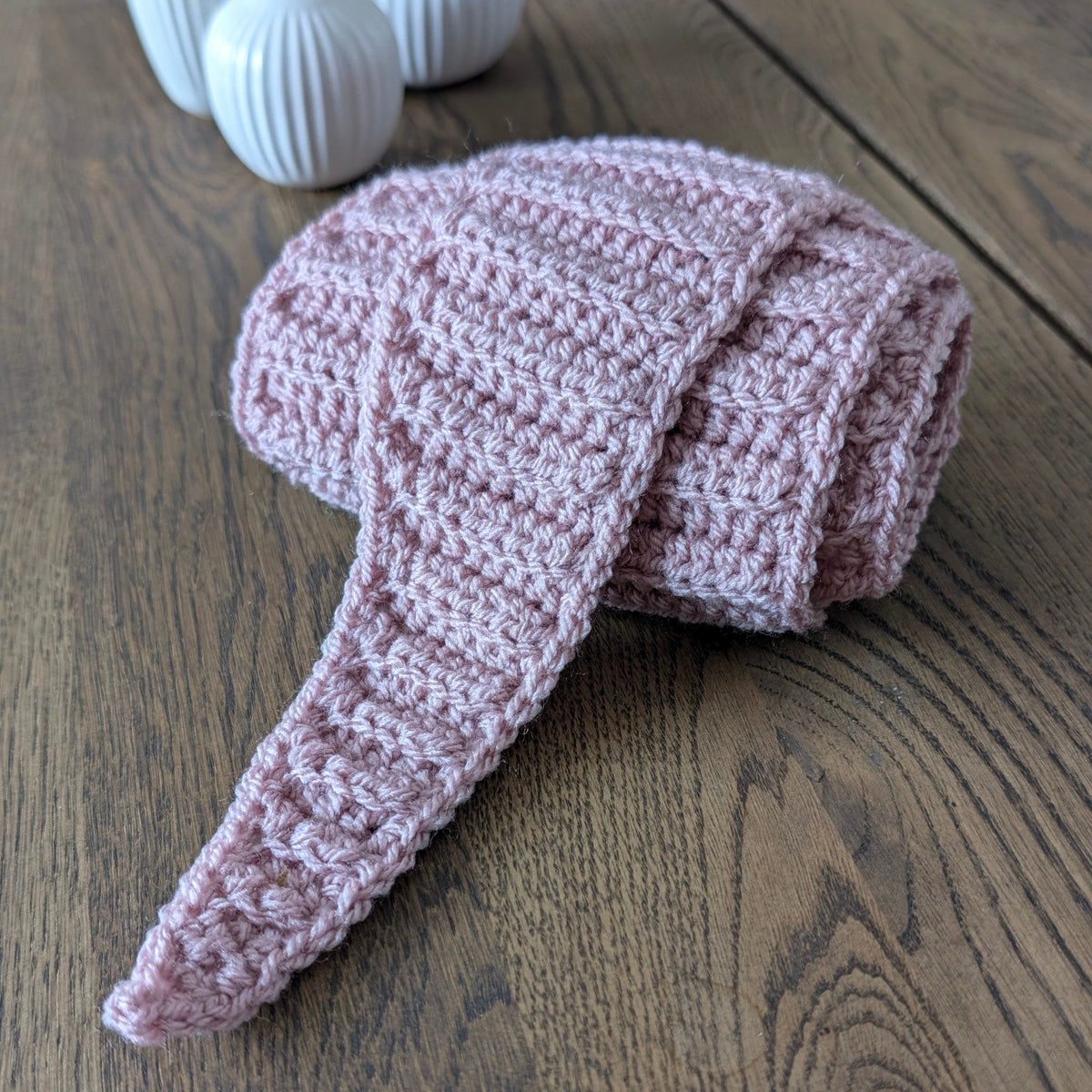 Folded dusty pink ribbed crochet scarf on a wooden table, showing its textured stitches and one long tapered pointed tip.