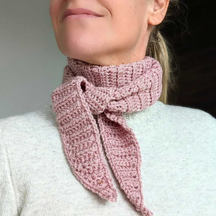 Side view of a woman in a cream knit sweater wearing a pink ribbed crochet neck scarf with the tapered ends tied loosely at the front.
