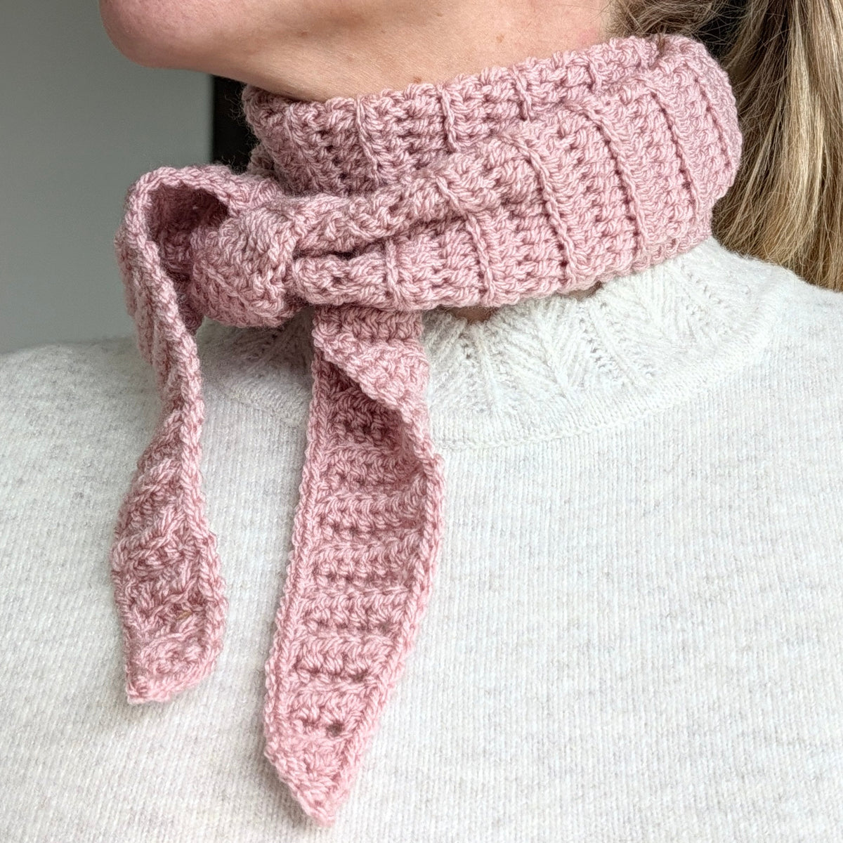 Woman wearing a dusty pink ribbed Sophie Scarf tied around her neck over a cream sweater, with two tapered crochet ends hanging in front.