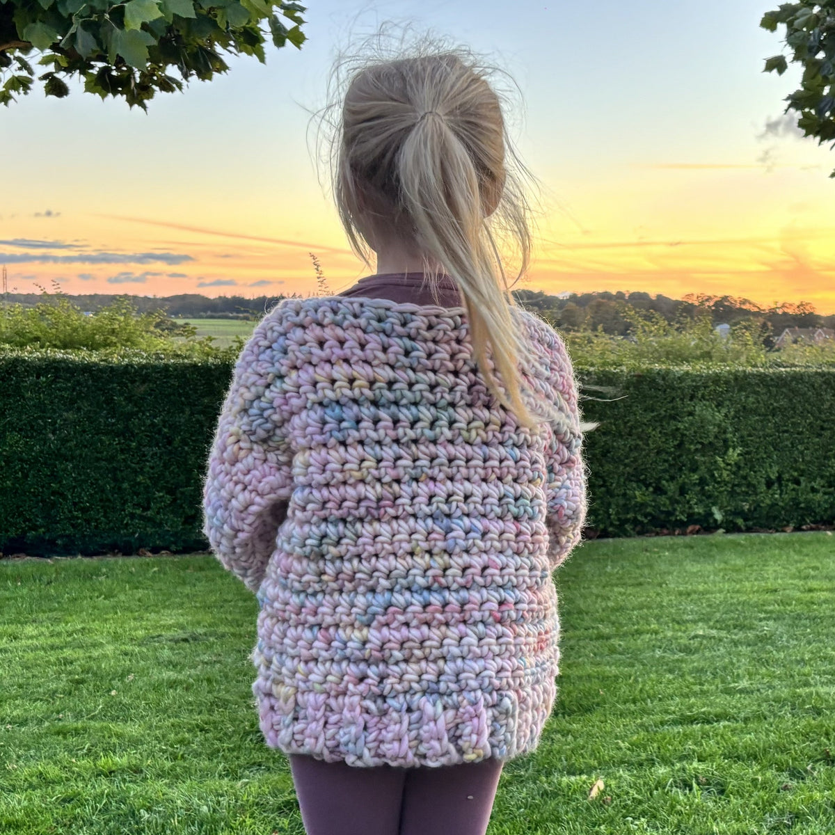 Back view of a handmade chunky crochet kids sweater in pastel pink, blue, lilac and mint, with textured stitches and a ribbed lower edge.